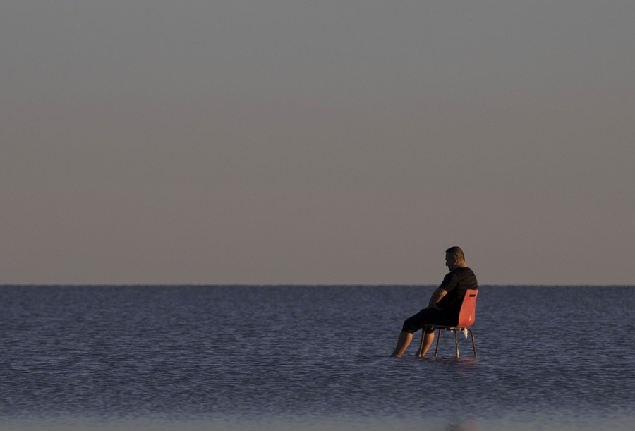 A person sits on a chair in shallow water at the edge of a vast lake.