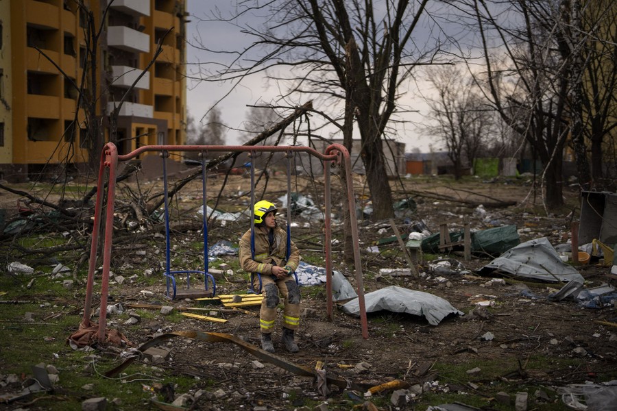 A firefighter sits on a swing set, amid debris, next to a building destroyed by a missile.