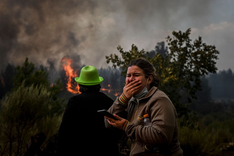 Two people watch an approaching wildfire as it burns through trees and brush.