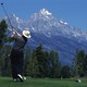 A golfer swings on a lush golf course with snow-capped mountains in the distance.