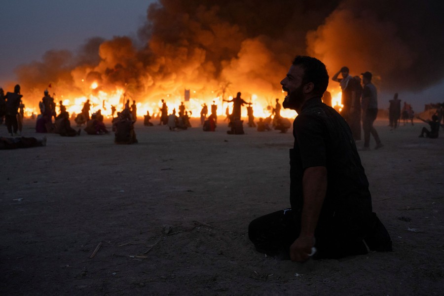 A man kneels on the ground, his mouth open, in front of a fire and a re-enactment of an ancient battle.