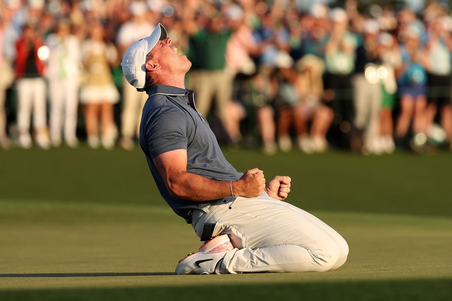 A golfer shouts in celebration, dropped to his knees, on a green, with a crowd cheering in the background.