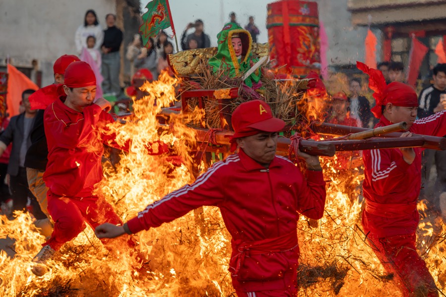 Four or five people carry a frame on their shoulders, holding up a small statue of a god, as they leap through a bonfire during a festival.