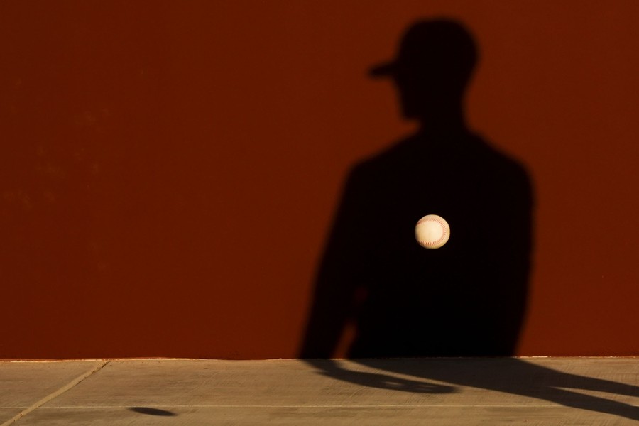 The shadow of a baseball player against a red wall. A baseball passes by just in front of the shadow.