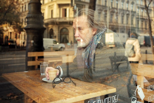 a portrait of Rod Dreher, wearing a gray coat and large scarf, sitting at a cafe table in Budapest