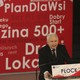 Jaroslaw Kaczyński, the leader of Poland's governing party, stands at a lectern in front of a banner with his party's policies.