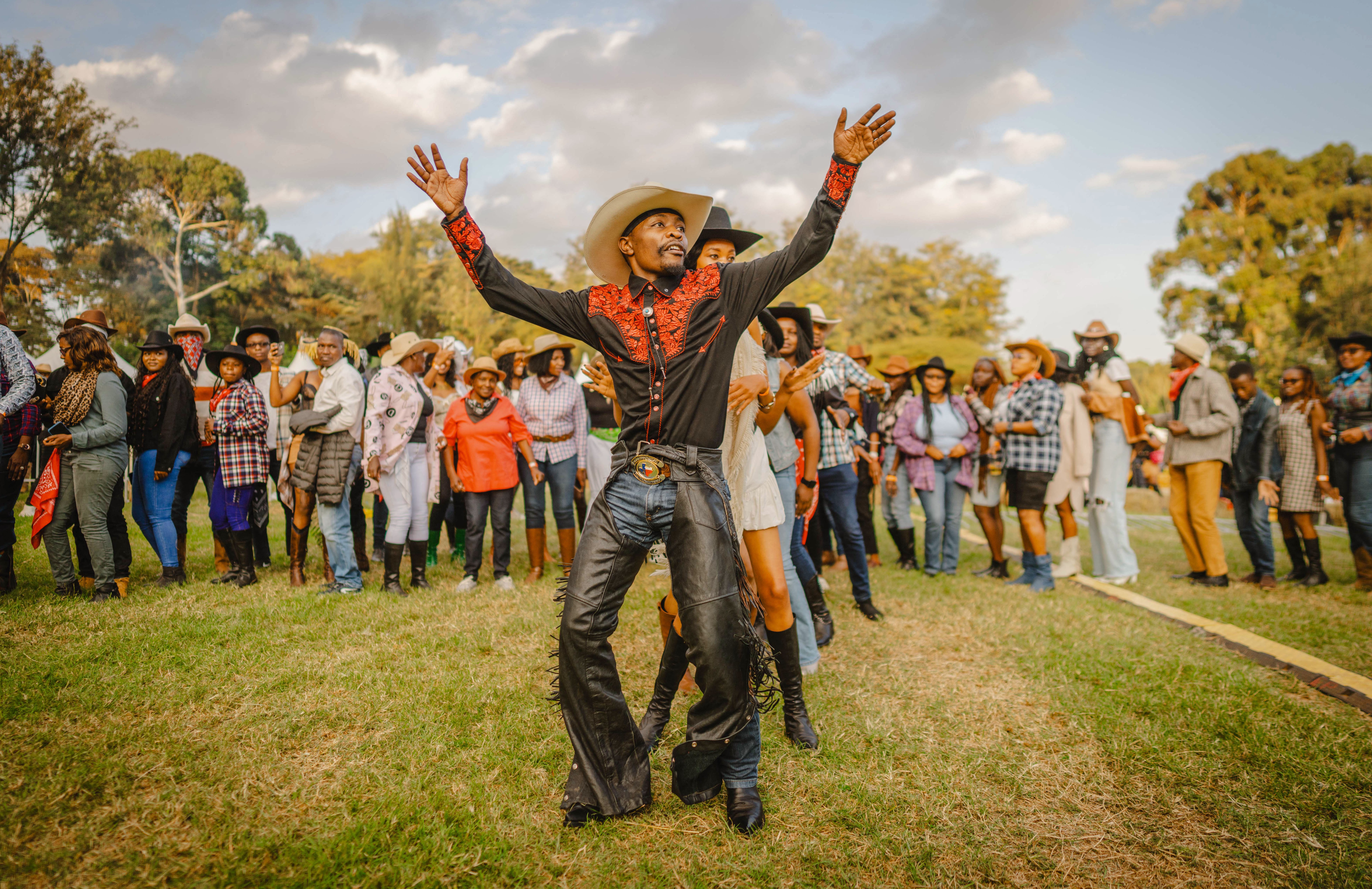 A man wearing chaps and a cowboy hat leads a line of dancing people at an outdoor music festival.