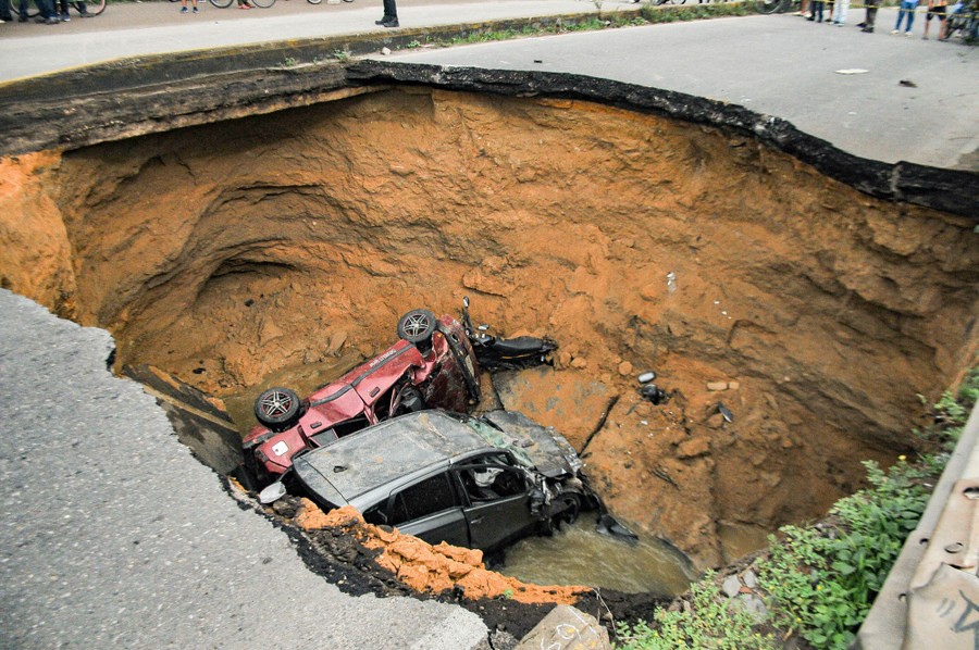 Several damaged cars sit in a deep hole below broken pavement.