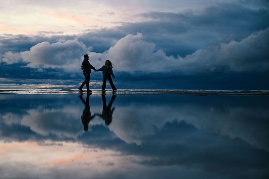 A couple taking a walk on a cloudy day are reflected in still water.