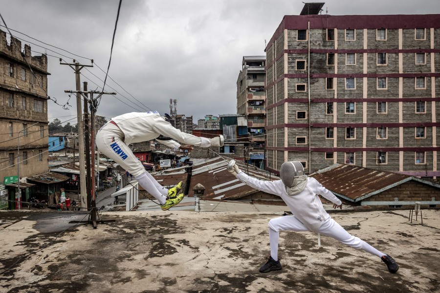 Two people wearing fencer's gear spar on a rooftop in a city setting.