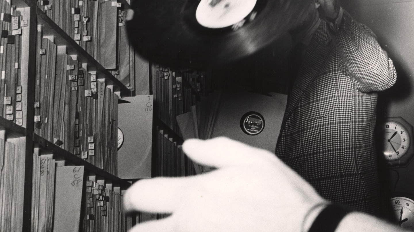 A black-and-white photo of a man in a plaid suit holding up a record, his face obscured