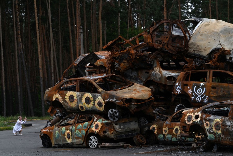 A woman takes a picture of a pile of destroyed vehicles, some painted with sunflowers.