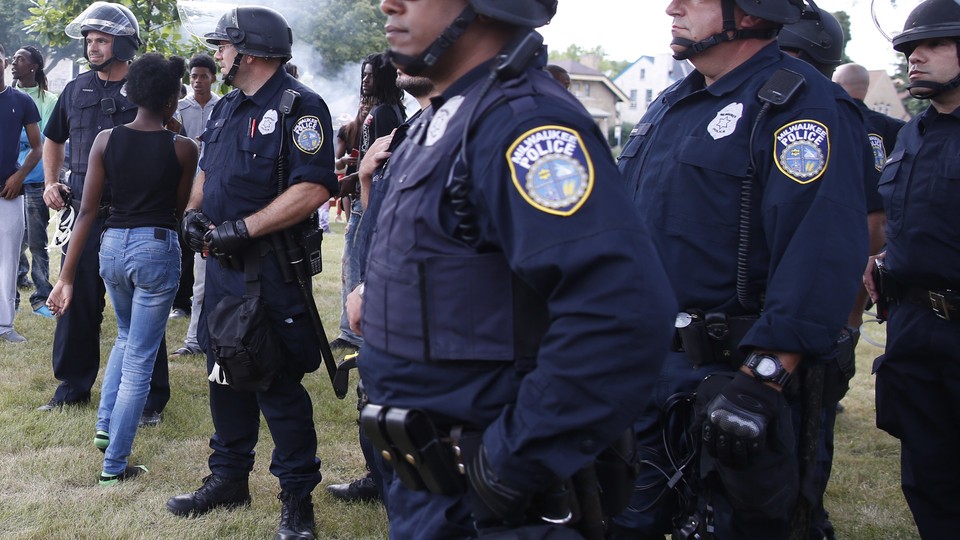 Five male police officers in full uniform and helmets stand looking toward the left; a young black woman walks through two of them with her back turned to the camera.