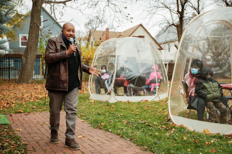 A man with a microphone walks while speaking to people gathered nearby in clear plastic tents.