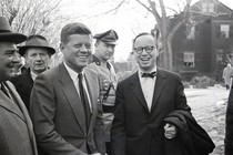 A black-and-white photograph of JFK and his biographer Arthur Schlesinger Jr. laughing in Boston winter, surrounded by a crowd of men