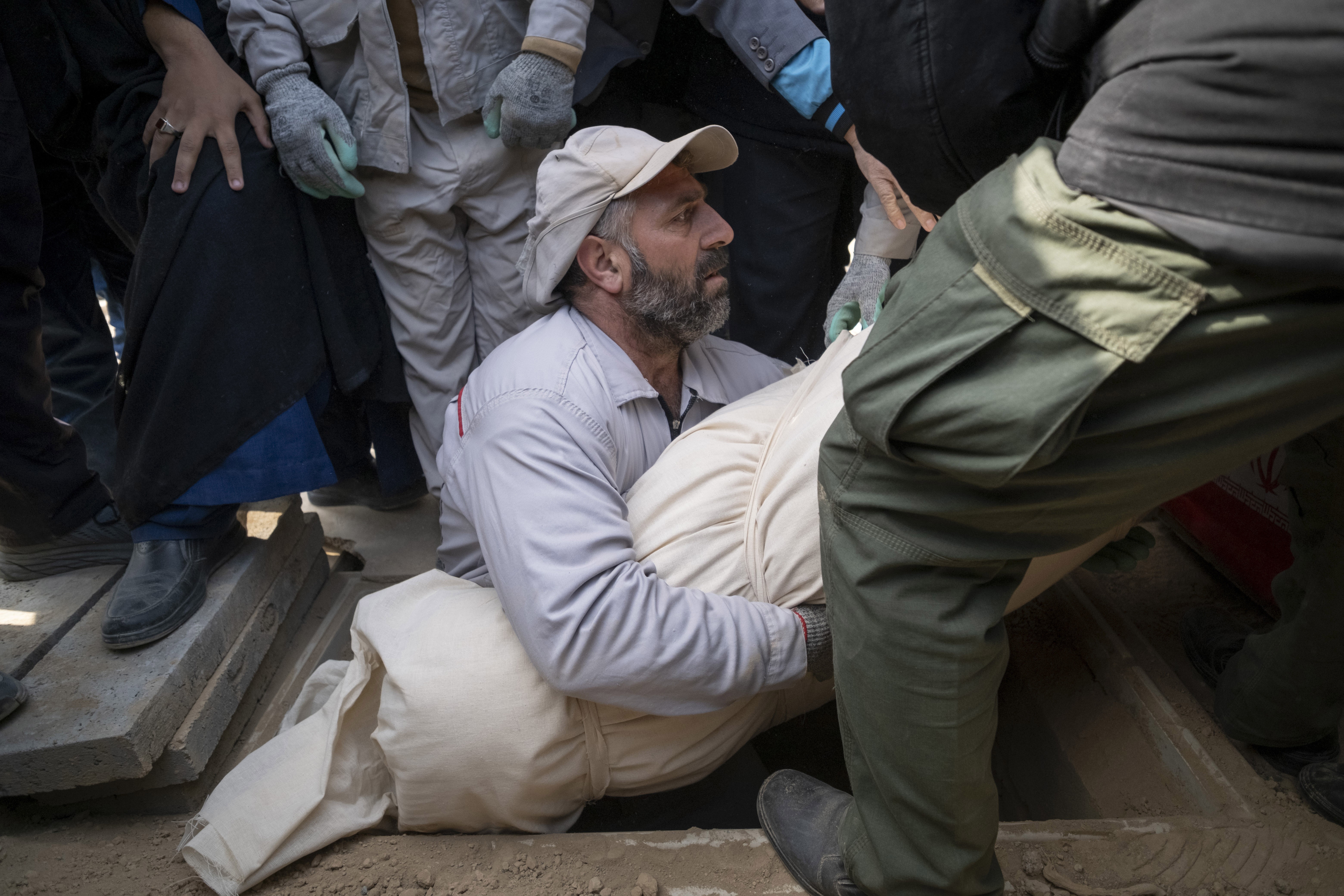 A man holds a shrouded body, lowering it into a grave, surrounded by other people.