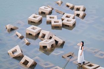 A woman stands on a cube-shaped concrete block, among many such blocks piled haphazardly in shallow water.