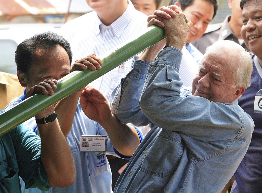 Jimmy Carter helps another person to pull down on a long bar, while making bricks.