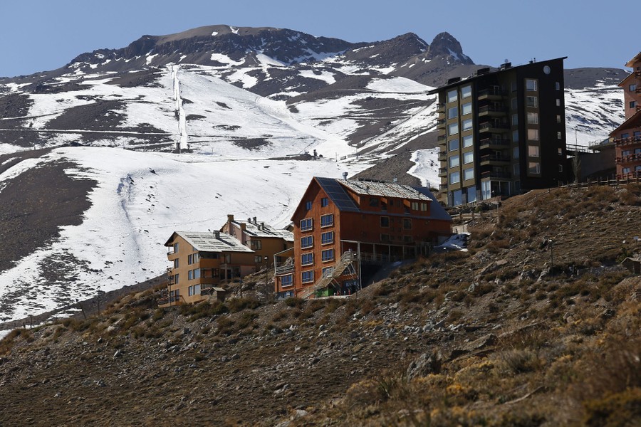 Ski resort buildings stand on a dry hillside near sparse patches of snow on upper slopes.