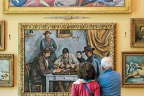 photo of the backs of two people looking at a wall of symmetrically arranged paintings including Seurat's "Models" and Cézanne's "The Card Players"