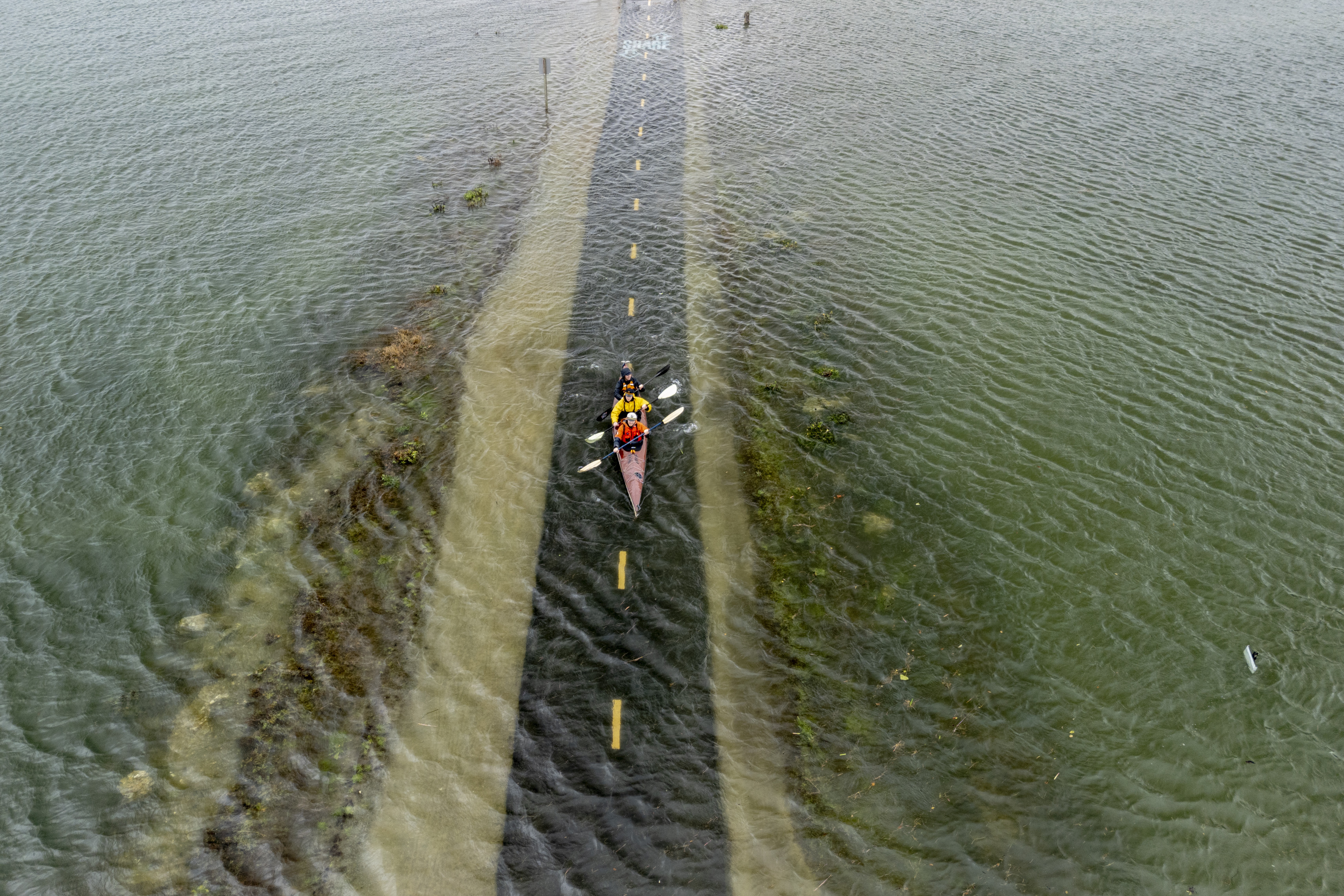 People in a three-person kayak paddle on shallow floodwater, showing a bike path just beneath the surface.