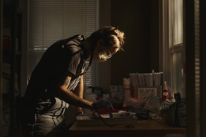 A medic leans over a desk in a darkened room, preparing medical supplies.