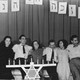In a black and white photo, a group of men and women laugh and smile in a row standing behind a menorah.
