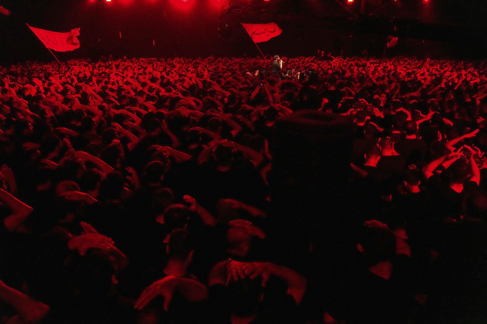 Dozens of men gather in a room beneath red light, performing a religious ceremony in Iran.