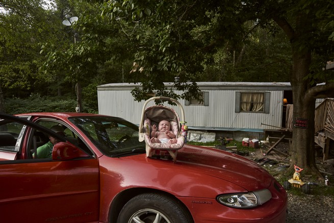 a baby sits in a car seat on the hood of a red car in front of a trailer home.
