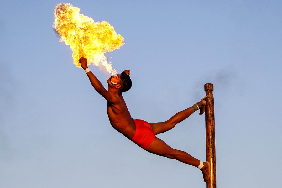A man stands against the side of a pole, with one foot tied near the top, stretching out sideways and breathing fire.