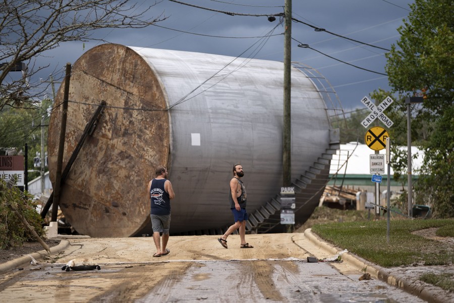 Two men stand on a road beside a large storage tank that has been toppled into the road by flooding.