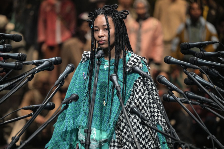 A model displays a draped garment, standing before a decorative collection of dozens of microphones.