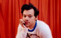 Harry Styles in a white sweater, blue chunky necklace, against orange-red curtain backdrop