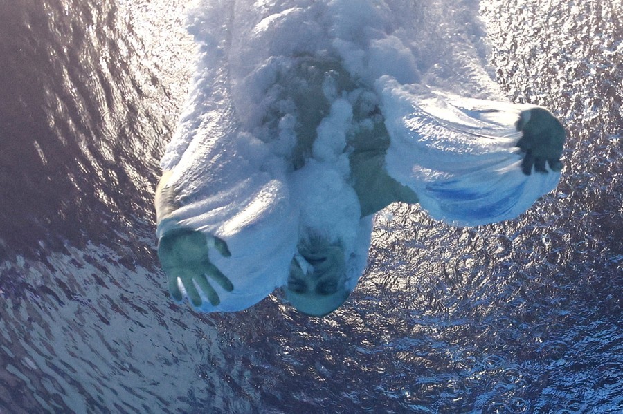 Captured from underwater, a diver enters a swimming pool, creating streams of bubbles with their head and hands.