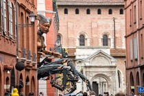 A giant mechanical puppet of a mythical human-scorpion creature moves through a narrow street.