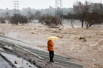 Photo of a person holding an umbrella beside the Los Angeles River during heavy rains