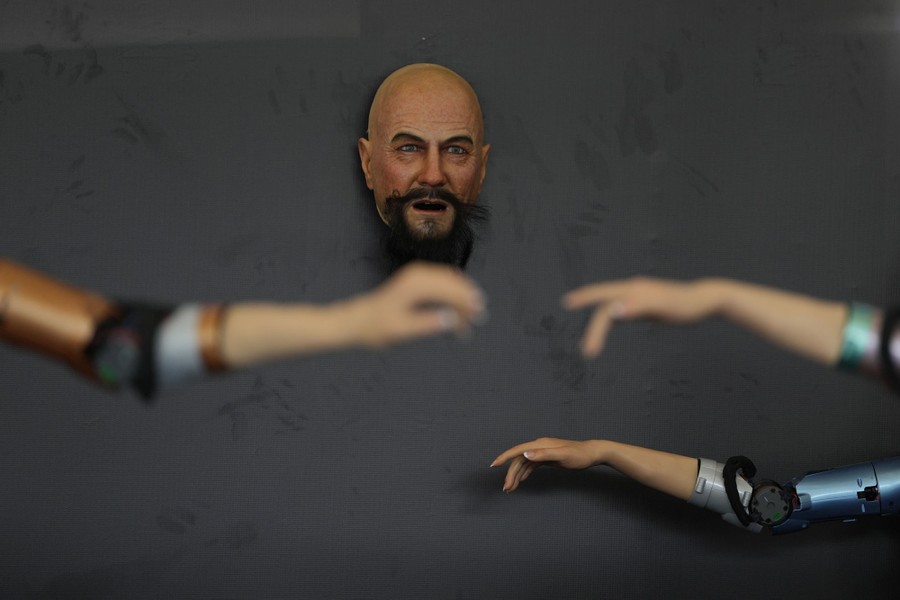 An artificial human face is attached to a wall, beyond the outstretched arms of several robotic devices.