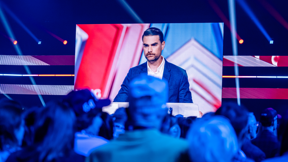 A giant television screen displays Ben Shapiro talking at a conservative conference, as viewers watch in the foreground.
