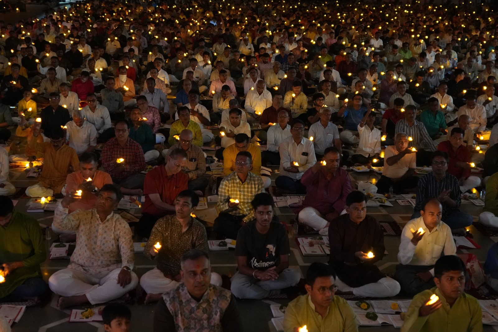 A large group of people sit together in rows, each holding a small oil lamp or candle.