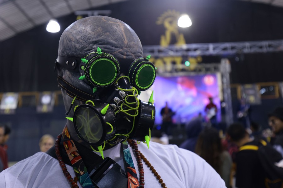 A tattooed person wears stylized goggles and gas mask inside a convention space.
