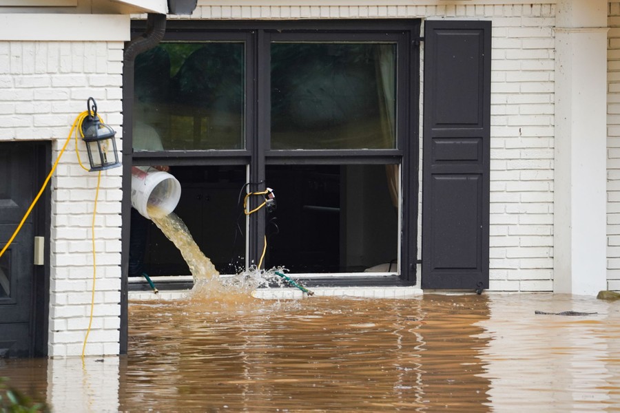A person dumps a bucketful of water out of a house's window, into floodwater outside.