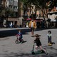 A man in a mask oversees a group of children playing on bikes and scooters in a plaza outside.