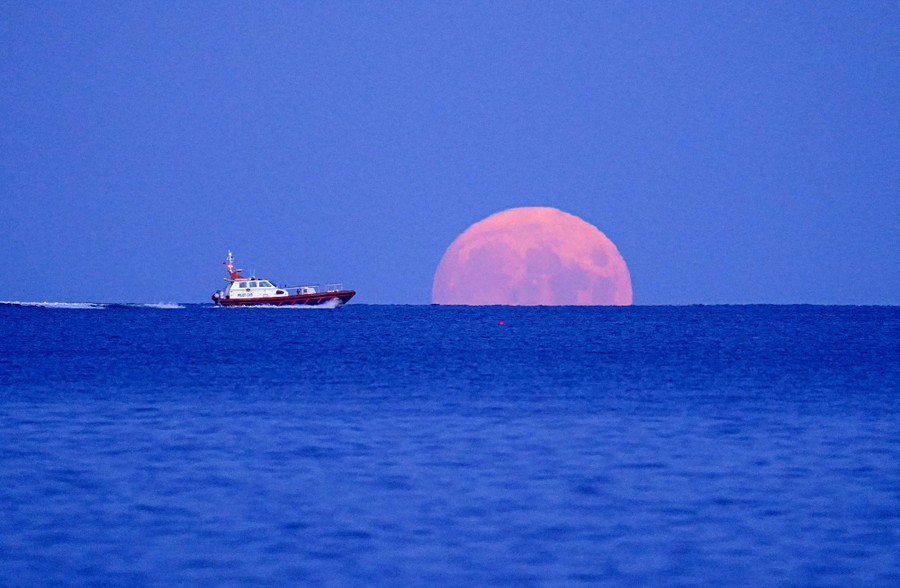 A boat passes by a rising full moon.