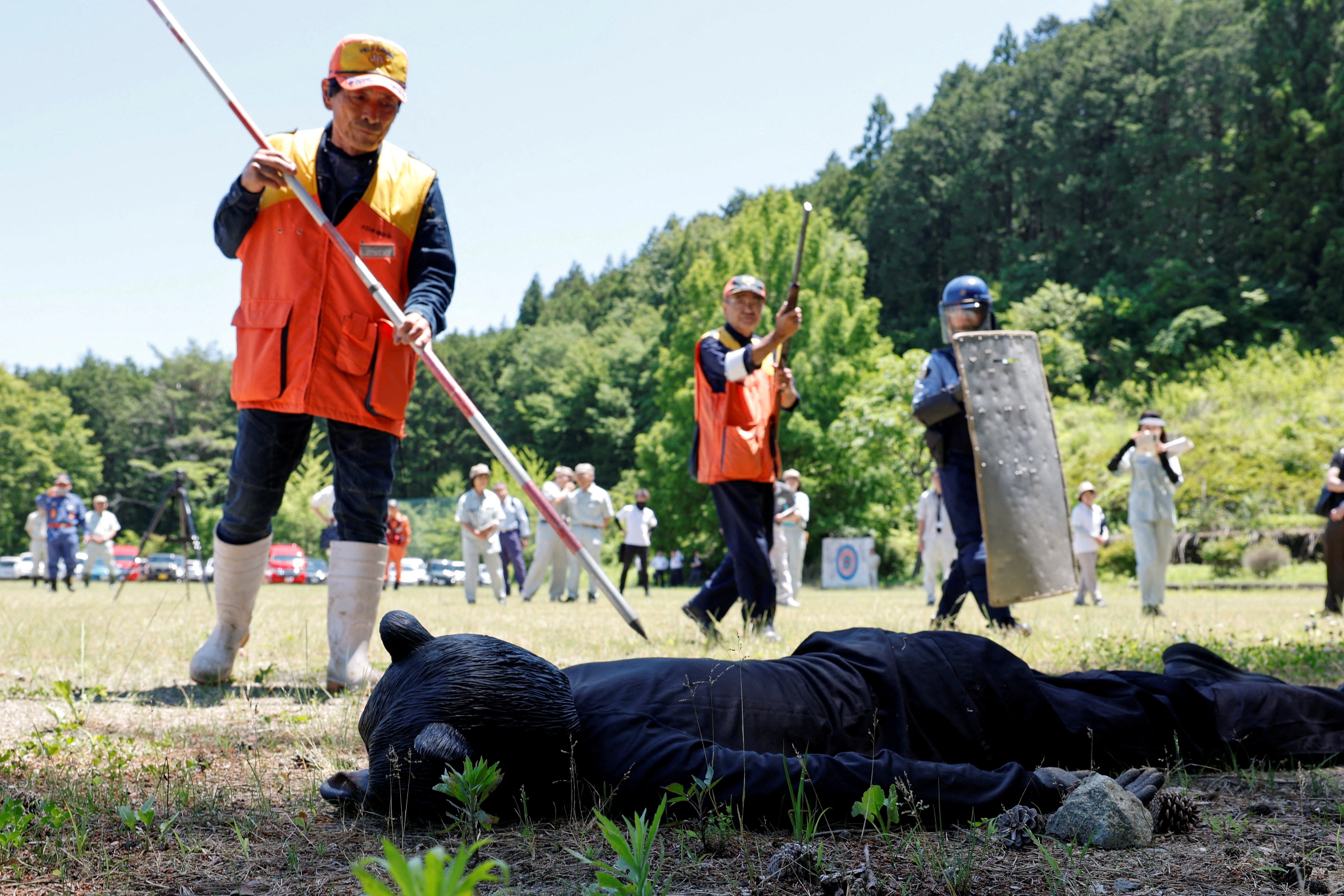 Two hunters and a police officer approach a man in a bear costume who pretends to be dead.