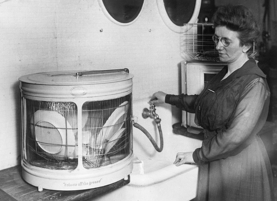 A woman operates a faucet at a kitchen sink, attached to a hose that leads to a cylindrical device that holds several plates, spraying them with water.