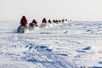 About a dozen people in red parkas ride snowmobiles in a line across a broad, flat expanse of snow and ice.