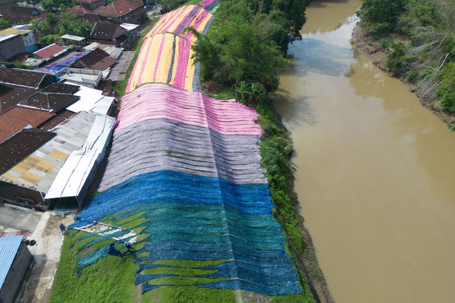An aerial view of many long strips of colored fabric laid out to dry on a berm alongside a river