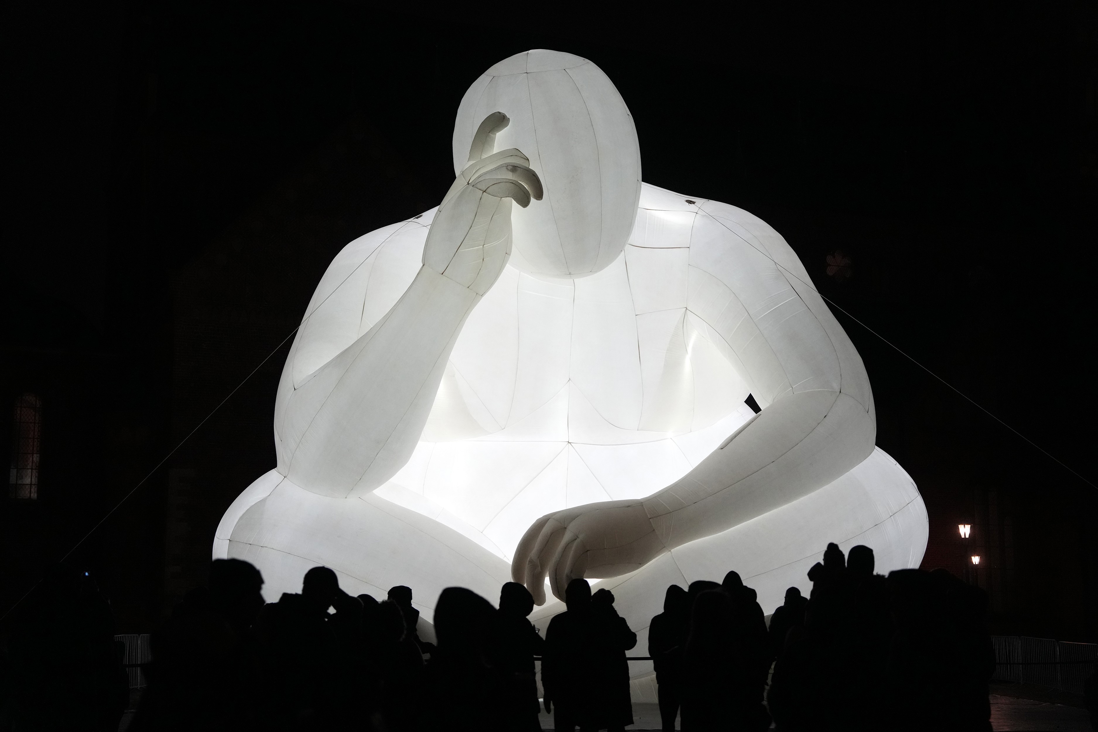 An illuminated inflatable structure shaped like a large person sitting cross-legged on the ground