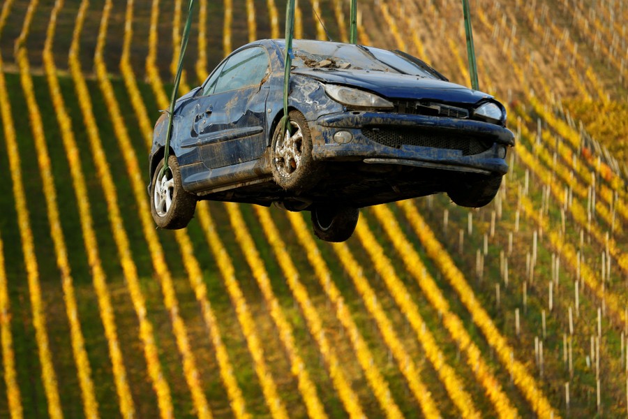 A wrecked car is lifted into the air by a crane, with a field in the background.