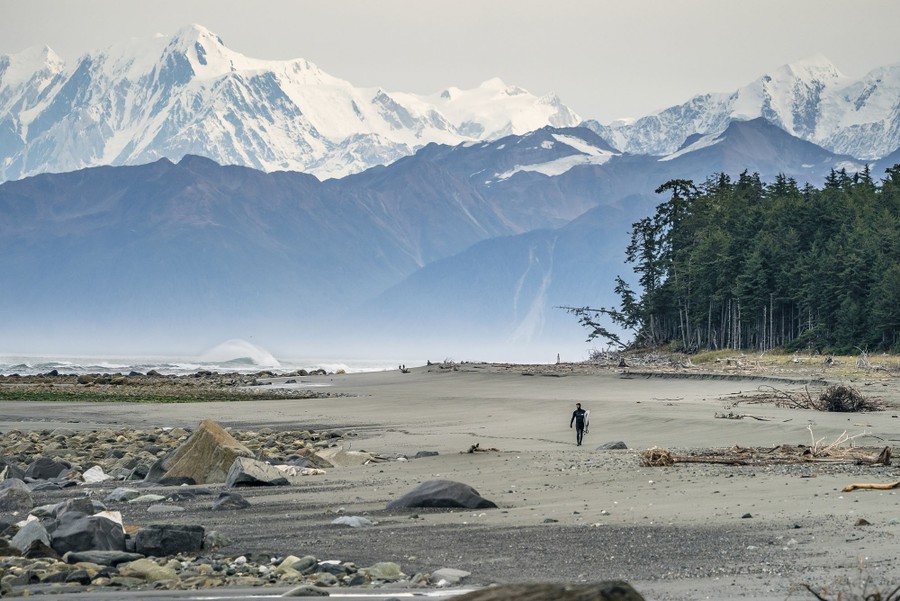 A surfer walks alone on a broad and somewhat rocky beach with tall, snowcapped mountains in the background.